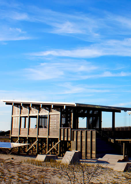 Modern beach house with slatted wooden exteriors under a clear blue sky, elevated on stilts above sandy terrain