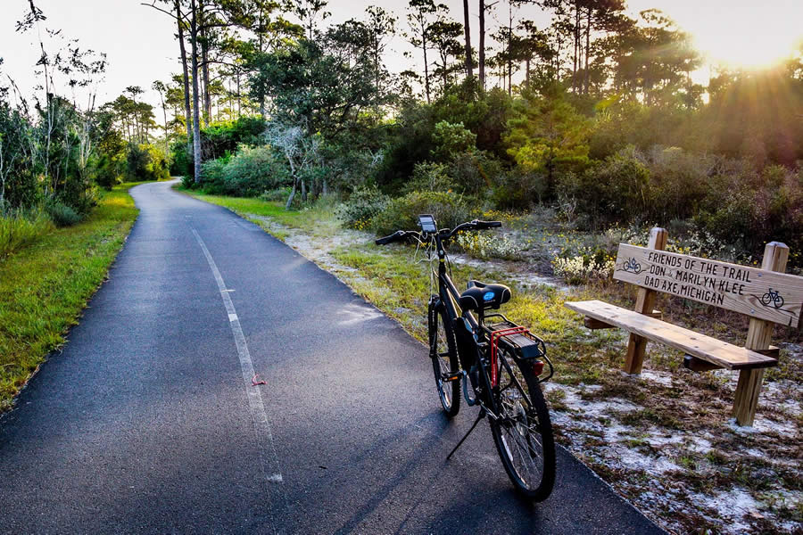 Bicycle parked beside a serene forest trail at sunset, with a bench dedicated to trail supporters