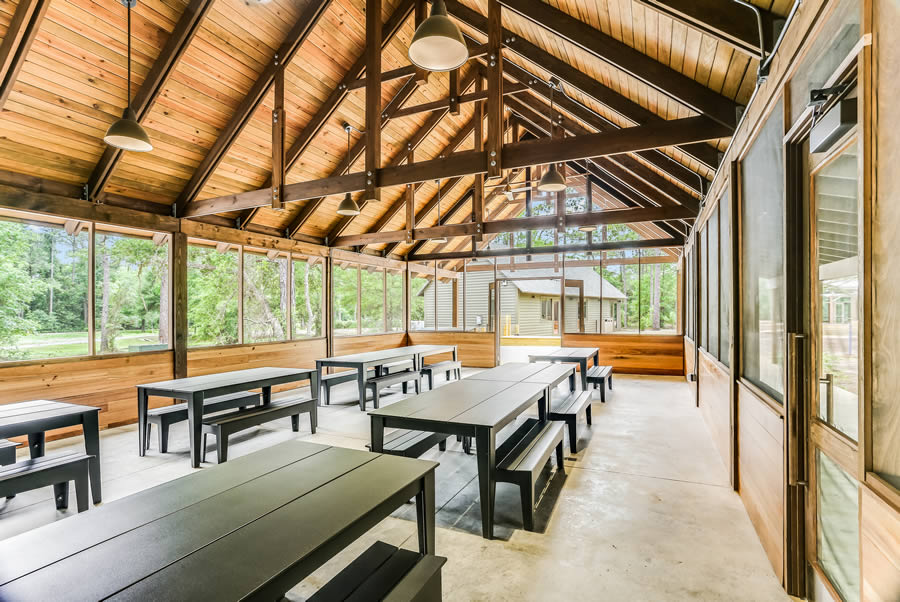 Spacious wooden dining hall with tables and benches, surrounded by large windows overlooking a forest